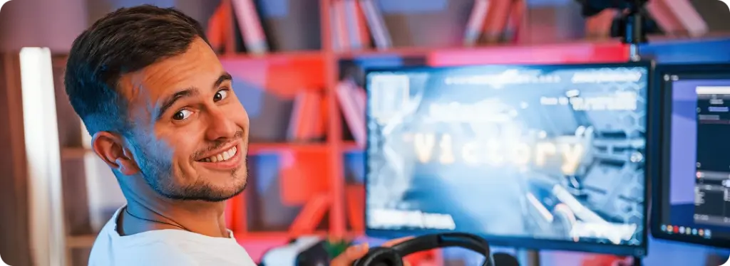 Een jonge man in een gamingopstelling, glimlachend en met een koptelefoon op. Op het scherm staat ‘overwinning’, wat doet vermoeden dat hij een spel heeft gewonnen. De kamer is sfeervol verlicht met rode en blauwe accenten