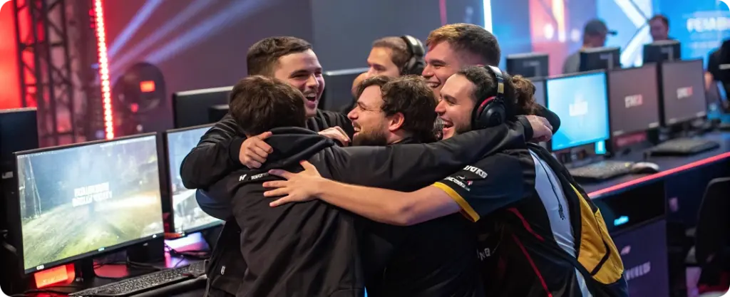Een groep esports-spelers in teamshirts viert het met een groepsomhelzing voor hun computerschermen. Het tafereel straalt enthousiasme en teamgeest uit