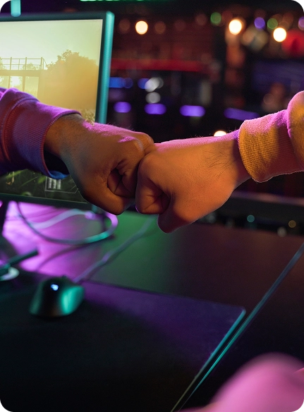 Twee mensen in kleurrijke sweatshirts geven elkaar een vuistje voor gamemonitors tegen een warm verlichte achtergrond, wat een gevoel van teamwork en kameraadschap uitstraalt